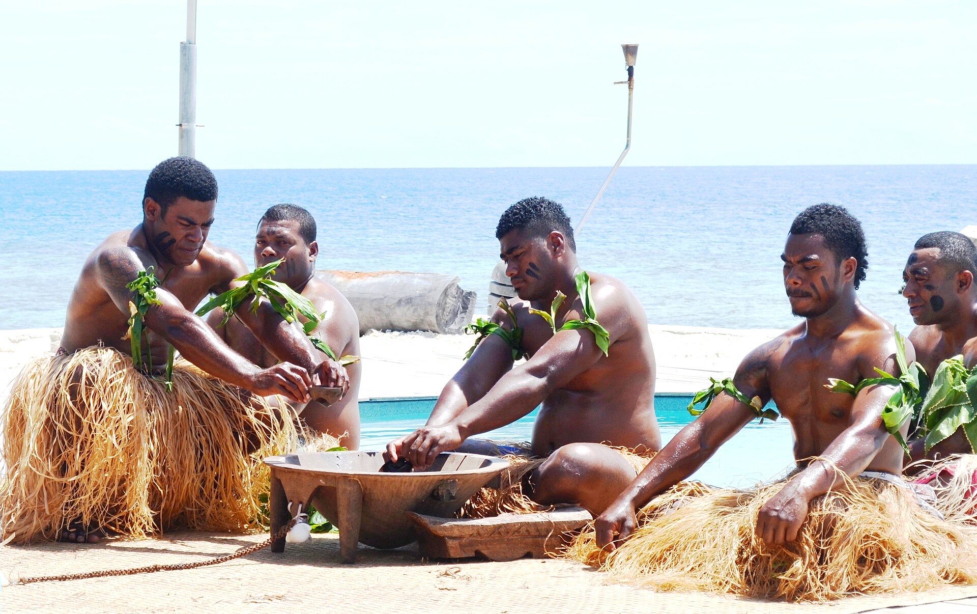 Fijian men in traditional dress preparing kava around a wooden tanoa bowl beside the sea